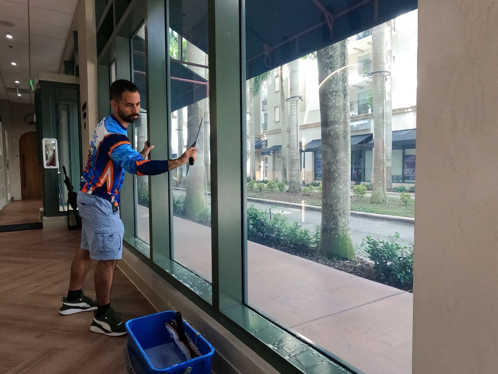 Shiny Windows technician squeegeeing the interior glass of a luxury medical aesthetic clinic at Shops at Merrick Park in Coral Gables, Florida, with a blue cleaning bucket on the floor beside him
