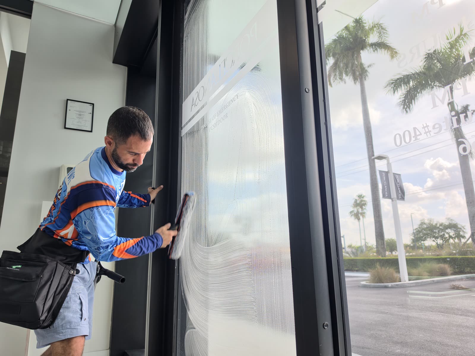 Shiny Windows technician in a branded blue and orange jersey using a squeegee to clean the tall exterior storefront glass entry doors at the Porcelanosa showroom in Doral, Miami, Florida, with reversed Porcelanosa signage visible on the glass and palm trees and the parking lot reflected behind him