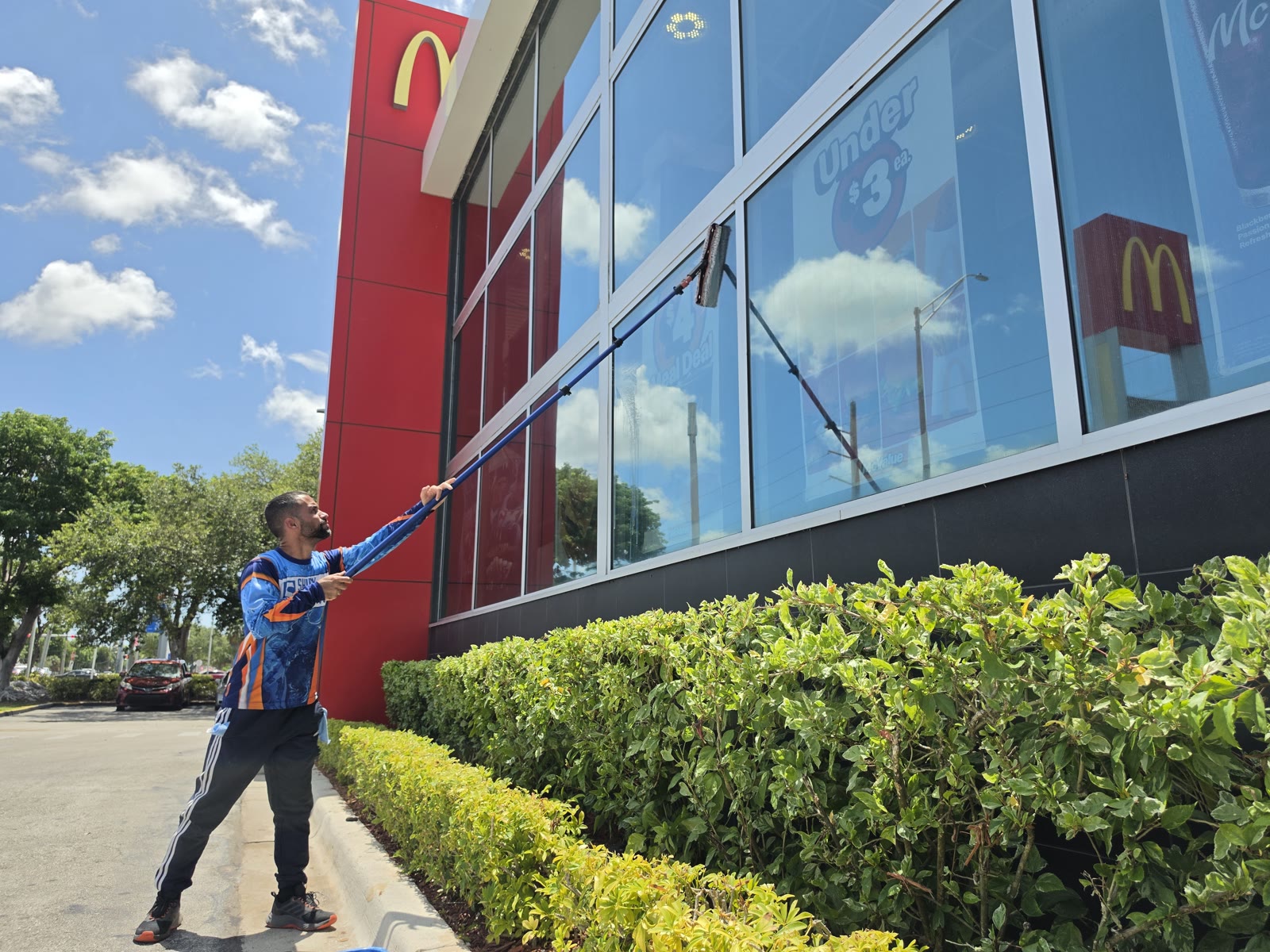 Shiny Windows technician in a branded blue and orange jersey using a Xero carbon fiber pole with a squeegee to clean the tall exterior storefront glass at the McDonald's in Country Walk, Kendall, Miami, Florida, with the red building and golden arches above, an Under $3 McCafe promo sign behind the glass, and green hedges along the walkway
