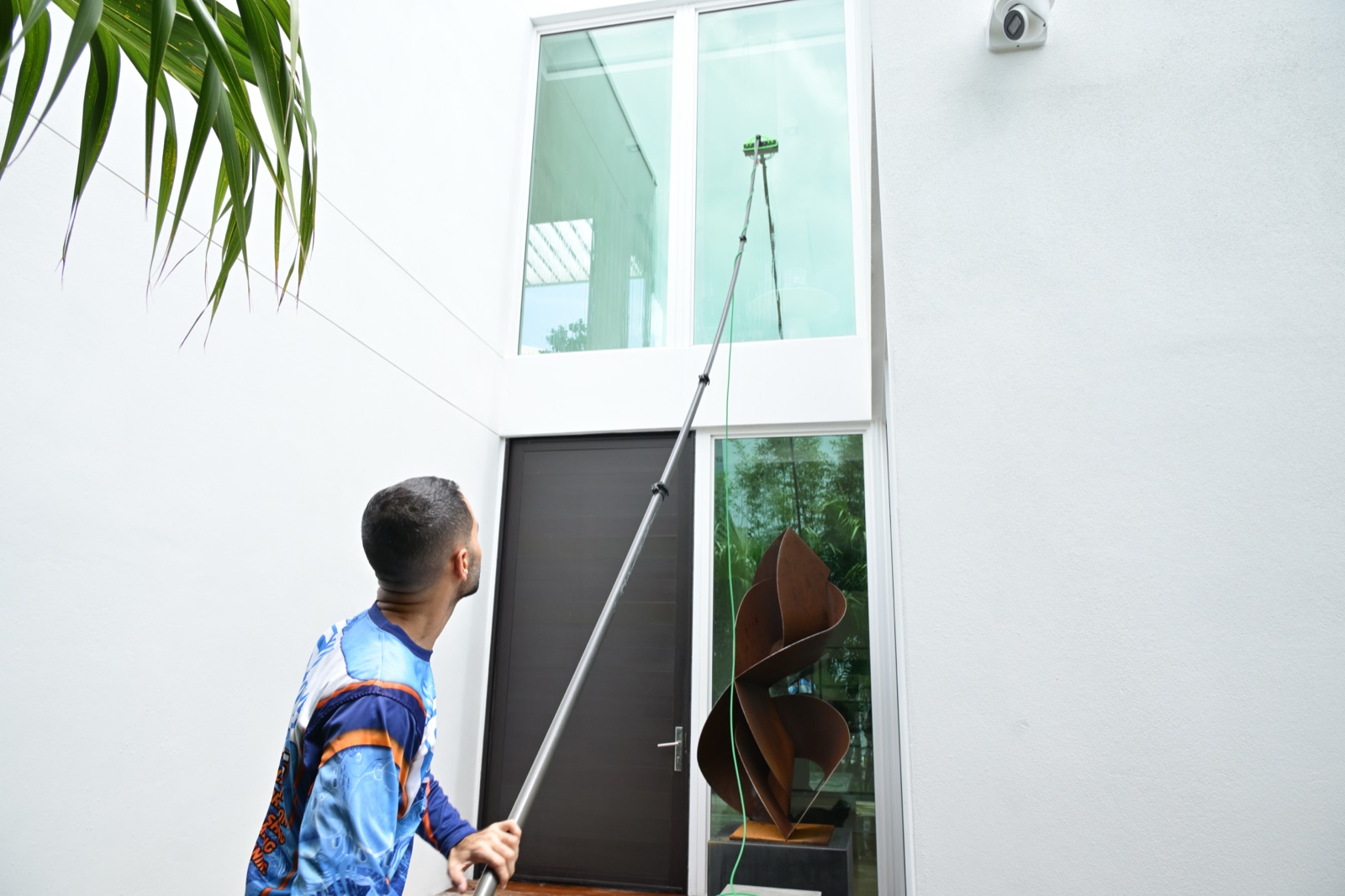 Shiny Windows technician using a water-fed pole to clean the tall second-story glass at a Ritz-Carlton Residences luxury home in Miami Beach, Florida, with a modern metal sculpture visible near the front entrance