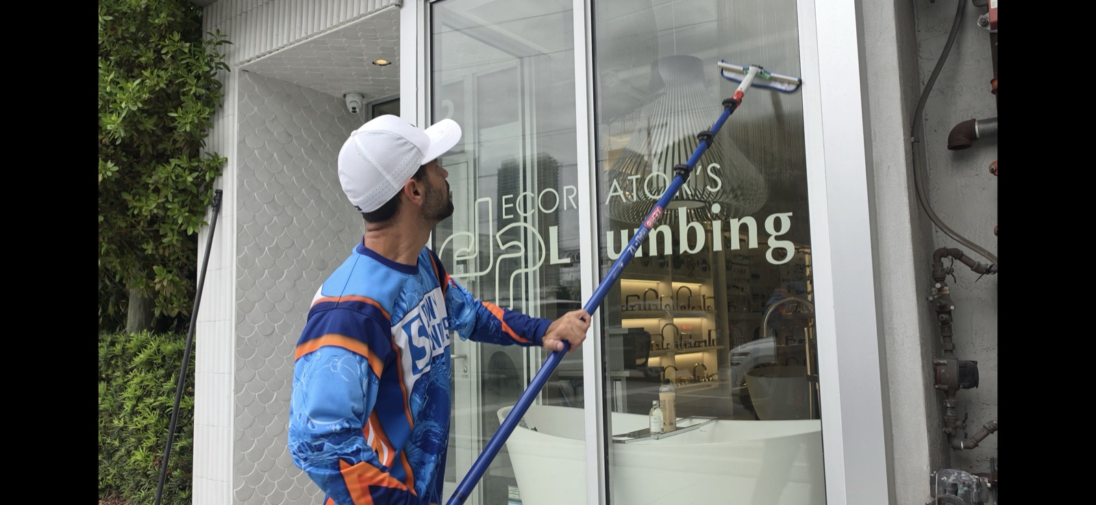 Shiny Windows technician in a branded blue and orange jersey using a pure-water-fed pole to clean the tall exterior glass storefront of Decorators Plumbing, a luxury kitchen and bath showroom in the Miami Design District, Miami, Florida, with green hedges at left and bath fixtures on display behind the glass