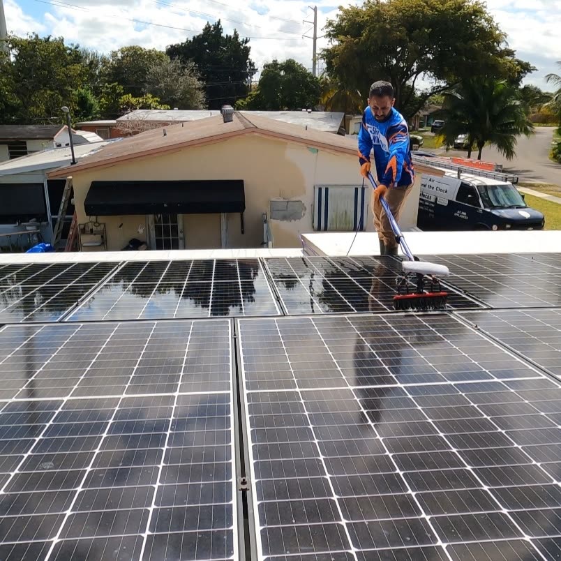 Shiny Windows technician cleaning rooftop residential solar panels in North Miami, Florida, using a soft-bristle brush on a water-fed pole, with the Shiny Windows service van visible in the driveway behind him