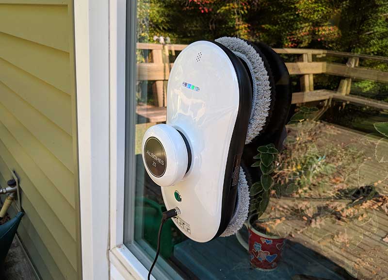Consumer window cleaning robot suctioned to an exterior window pane with its microfiber rollers and power cable visible, the kind of $200 Amazon robot most Miami homeowners buy before realizing it can't replace a professional window cleaning service